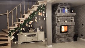 The living room has a fireplace with burning wood, a wooden staircase with railing to the second floor and a dresser. The staircase is decorated for Christmas and New Year with fir garland and lights - Powered by Shutterstock - Get 15% off with code: PIKWIZARD15