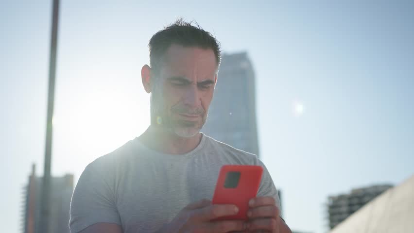 Mature Caucasian muscular man standing concentrated using mobile phone. Male in sports wear looking serious cell doing scrolling outdoor. Healthy people connected in digital display. Lens flare 