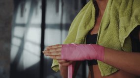 Woman wrapping her hands with pink boxing wraps, with towel draped over her shoulder, getting ready for her training session in gritty gym environment - Powered by Shutterstock - Get 15% off with code: PIKWIZARD15