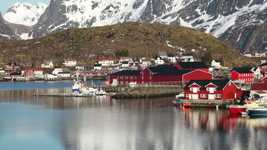Small traditional Scandinavian village with red houses on the picturesque fjord coast. Beautiful mountains covered white snow. Lofoten Island, Norway. Cottage on the shore of the fjord.