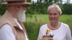 A stylish gray-bearded elderly husband in sunglasses and a brimmed hat gives his elderly wife a taste of his ice cream cone, while standing outdoors in a city park in summer. A married couple tasting - Powered by Shutterstock - Get 15% off with code: PIKWIZARD15