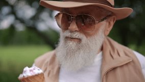 Close-up. Stylish bearded gray-haired elderly man in sunglasses and a brimmed hat got dirty, eating ice cream in a cone while standing outdoors in the summer in a park. An older woman wipes the - Powered by Shutterstock - Get 15% off with code: PIKWIZARD15