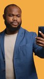 Vertical video Joyous man using smartphone to take selfies and post them on social media. Happy BIPOC person taking photos using phone selfie camera, isolated over yellow studio background, camera A