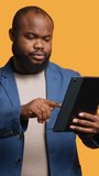 Vertical video Man happily scrolling on tablet touchscreen to check social media feed. African american person browsing internet websites, entertained by content, studio background, camera A