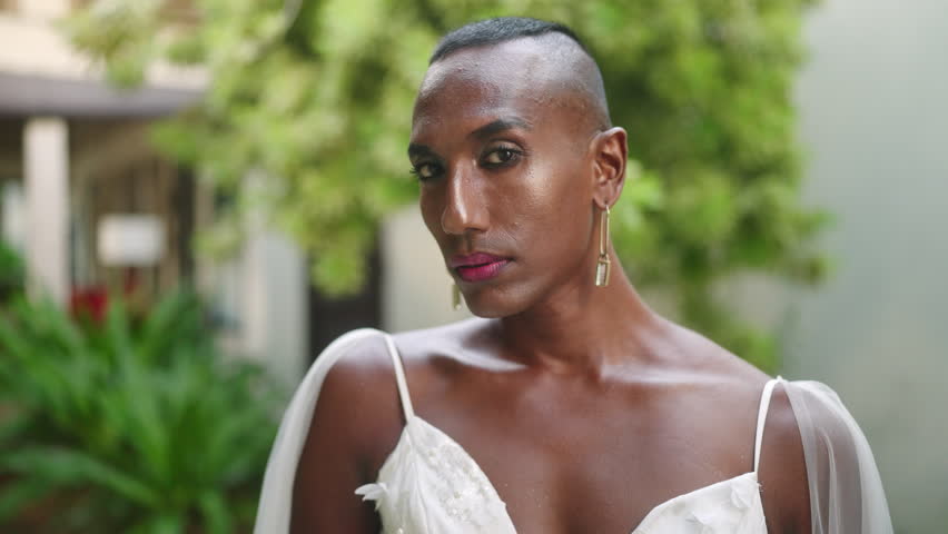 Gay black person poses in wedding dress, brass earrings, jewelry. Non traditional bride shows beauty, hands with a neat manicure, make-up, bracelet with gem. LGBTQ fashion shoot in villa green garden.