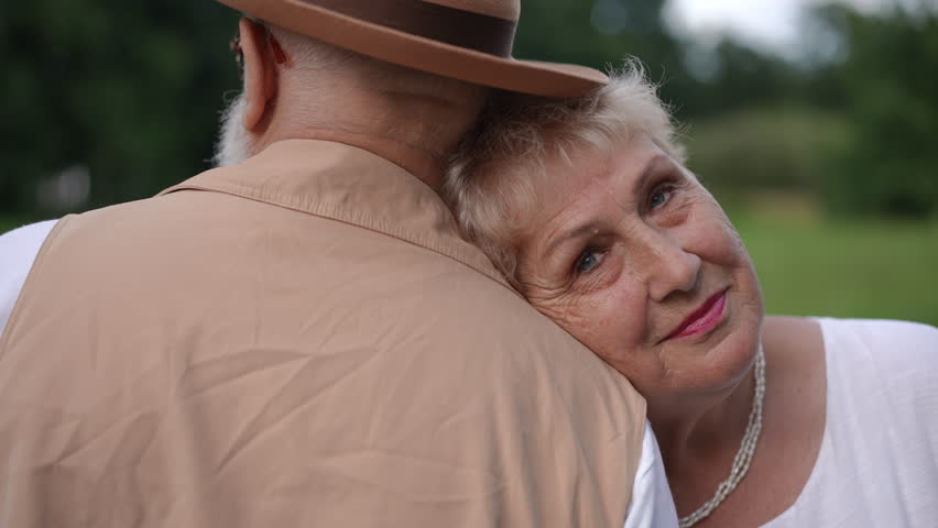 Close-up. The head of the elderly wife rests on the shoulder of her elderly husband, who stands with his back to the camera. An older woman looks at the camera with a smile