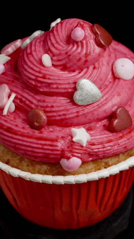 Vertical video. A red cupcake with frosting decorated with hearts is spinning, and a male hand picks it up. On a black background.