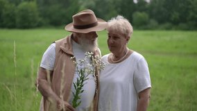 In the meadow, an elderly gray-bearded man in a brimmed hat gives an elderly woman meadow flowers. An elderly couple is talking to each other and smilingly looking at the flower in hand - Powered by Shutterstock - Get 15% off with code: PIKWIZARD15