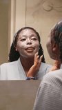 Skincare, Young Black African woman with long braids wearing blue bathrobe, standing in front of mirror in the bathroom, applying facemask to her skin. Vertical Video