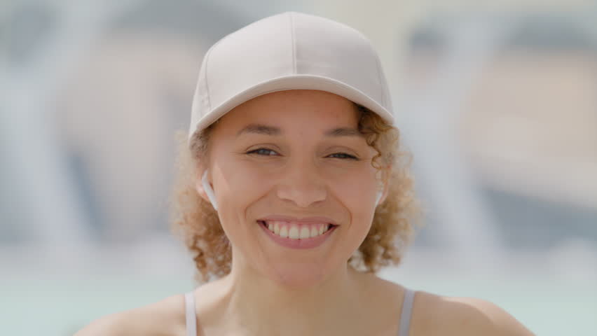 Radiant young woman with curly hair and a white cap, grinning broadly, enjoying a sunny day outdoors with a soft-focus background that implies a relaxed and joyful ambiance. Slow motion. 