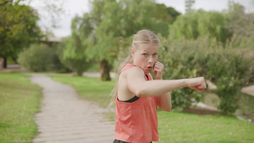 Young fit sport woman in a red sleeveless top boxing at park, Fight with shadow. Natural daylight and blurred greenery background accentuate the outdoor setting. Slow motion. 