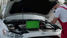 Mechanic repairs broken car under open hood laptop green chroma key screen nearby. Integration of digital visuals into video green chroma key presentation auto service meticulous work of mechanic.