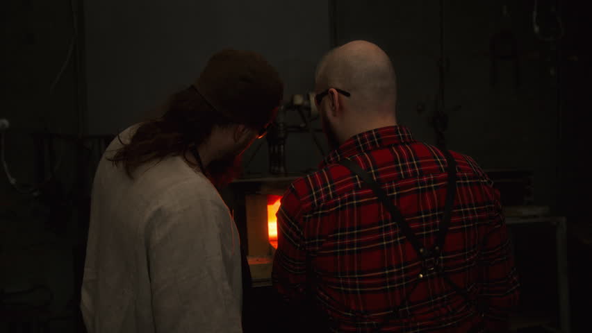 Two blacksmiths stay near a stone furnace in a forge, with a red flame flaring up. They push a metal product inside to heat and melt it