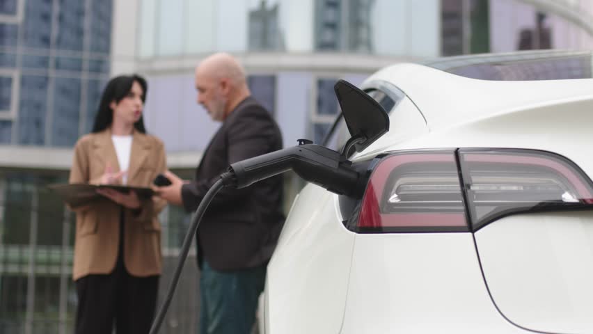 An unrecognizable modern electric car is charging. In the background, two business people, a man and a woman, sign a contract near a business center.
