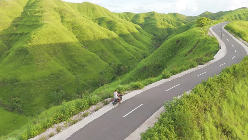 Sumba, Indonesia. A man is riding a motorcycle down a winding road with a lush green hillside in the background