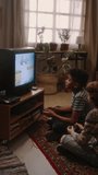 Vertical side handheld footage of two diverse boys sitting on floor in front of tube TV and playing old-school arcade video game at retro-styled home, 90s