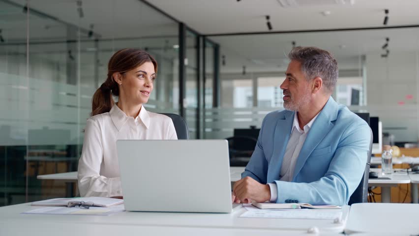 Two successful happy professional business partners executive leaders managers wearing suits working in office looking at computer technology discussing digital strategy together at corporate meeting.