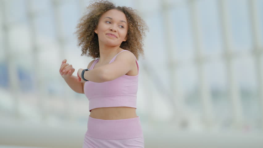 Athletic young woman in pink sportswear takes a moment to stretch her arms after an energizing outdoor workout, set against an urban architectural background. Slow motion.  Royalty-Free Stock Footage #3489389039