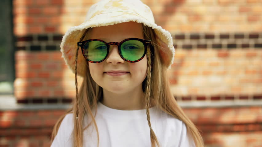 Portrait little stylish kid girl in summer sunglasses and a panama on city street. Child smiles and looks at camera.