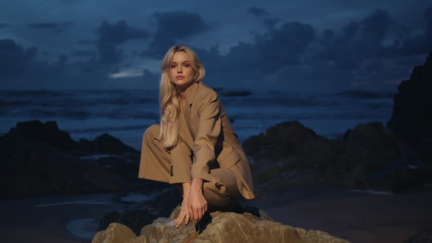 Sexy girl sitting rocks on night ocean island. Fashion model shooting dark coast looking camera in projector light. Elegant charming woman posing rocky beach at dusk. Beautiful seductive lady outdoors