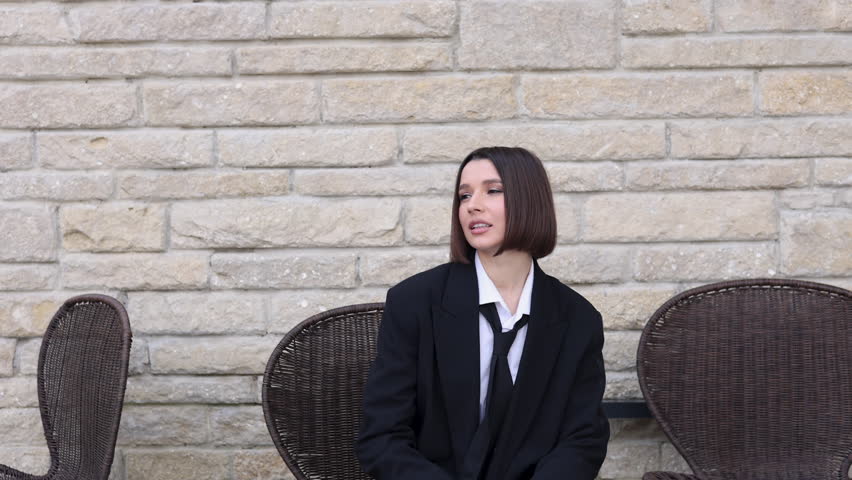 beautiful woman with short hair and business suit sitting on chair.girl doing brave moves with leg up.female in black jacket and white shirt with bag in hand,against brick wall