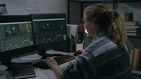 Medium side of young Caucasian female postproduction designer in casual shirt sitting at desk, editing raw footage in professional software on dual computer monitors, alone in office