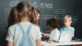 Girl smile and wave to camera while friend working or learning engineering code or prompt in STEM technology classroom. Student looking at camera and greeting while children using laptop. Erudition. - Powered by Shutterstock - Get 15% off with code: PIKWIZARD15
