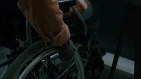 Medium close-up tilting shot of young black man with mobility impairment, using wheelchair, riding up towards desk, editing video clips for movie or film on computer with dual screen
