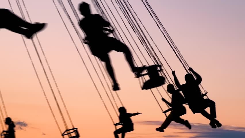 Unrecognizable silhouettes of people ride on the chained carousel against the warm evenning sky at sunset with at amusement park. Entertainment, summer, fun and holiday concept