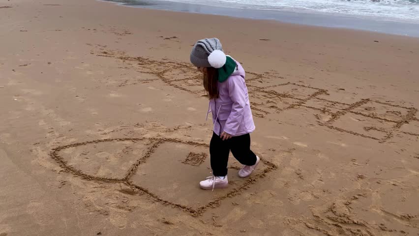 Toddler girl drawing on the sand on the beach with ocean view. Daily routine outdoor activities with family and kids. Playing outdoor games.