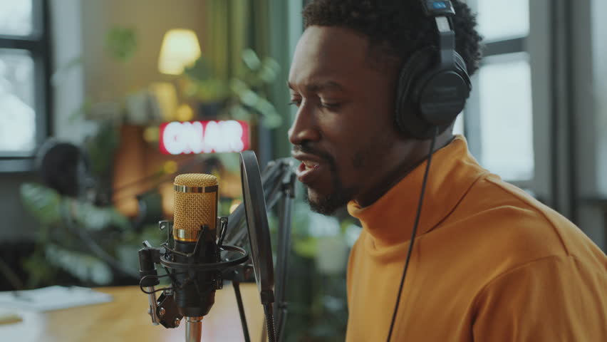 Young African American blogger in headphones talking in condenser microphone and recording new episode of audio podcast in studio