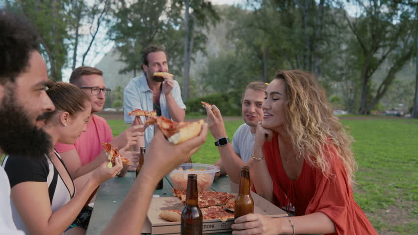 Group of relaxed friends laugh and share stories while enjoying beer and slices of pizza at an outdoor table in a lush park setting. Joy and ease are evident in this moment of carefree relaxation  Royalty-Free Stock Footage #3489702681