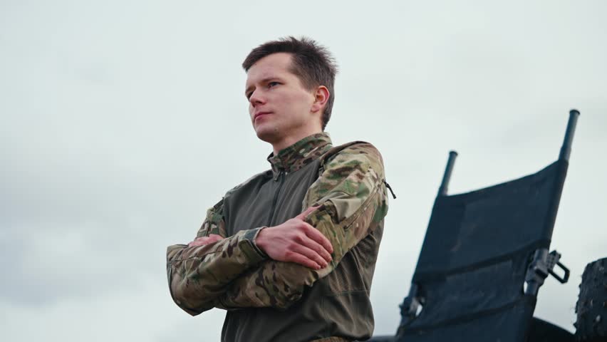 From the side a confident man with a short haircut in an army camouflage uniform stands confidently against the sky and looks to the side with his arms folded on his chest outside the city