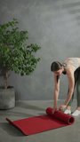 Vertical video of a happy young brunette girl in a black top and green pants lays out a red sports mat and starts her exercise at home in a modern apartment