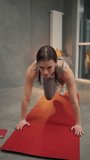 Close-up vertical video of a confident brunette girl in a sports summer uniform doing push-ups and sprints on a red sports mat in a sports mat in a modern apartment at home