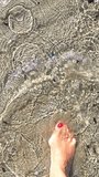 A person walks in the water with red-painted toenails. A beautiful and natural pattern has been formed in the sand. A woman walks in the shallow water on the beach in Thailand.
