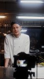 A man in a chef's uniform records the cooking process on video by putting bacon in a frying pan
