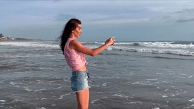 A beautiful girl takes pictures of the ocean on her smartphone. A woman walks along the coast of a popular beach and shoots a video of foaming sea waves at sunset for social media. . High quality 4k 