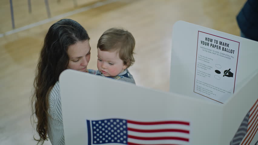 American citizens come to vote in polling station. Female voter with baby on hands stands at voting booth. Political races of US presidential candidates. National Election Day. Civic duty. Dolly shot.