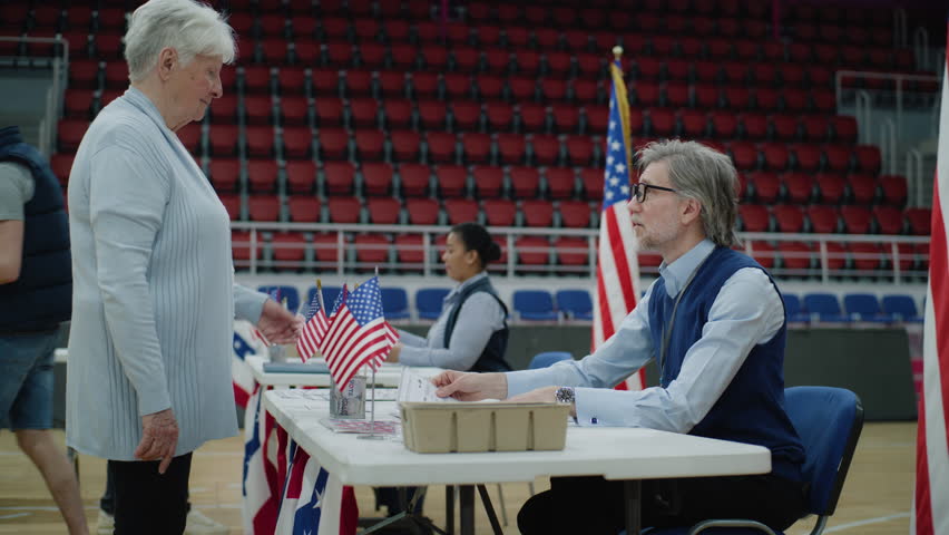Elderly woman talks with polling officer and takes vote bulletin. American citizens come to vote in polling station. Political races of US presidential candidates. National Election Day. Civic duty.