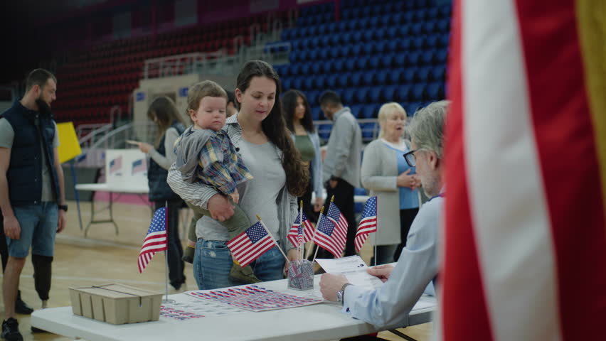 Female American citizen comes to vote in polling station. Woman with baby on hands talks with polling officer and takes bulletin. Political races of US presidential candidates. National Election Day. - Powered by Shutterstock - Get 15% off with code: PIKWIZARD15