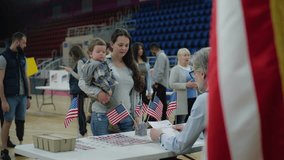 Female American citizen comes to vote in polling station. Woman with baby on hands talks with polling officer and takes bulletin. Political races of US presidential candidates. National Election Day. - Powered by Shutterstock - Get 15% off with code: PIKWIZARD15