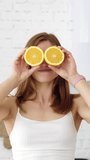 A cheerful girl covers her eyes with orange slices. Emotions in the white kitchen. Healthy eating. Vertical video.