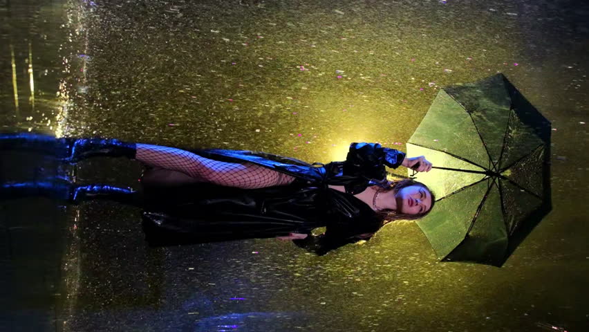 Young woman wearing lengthy black raincoat stands under rainfall. Torrent of water descends onto lady from above in dimly lit room