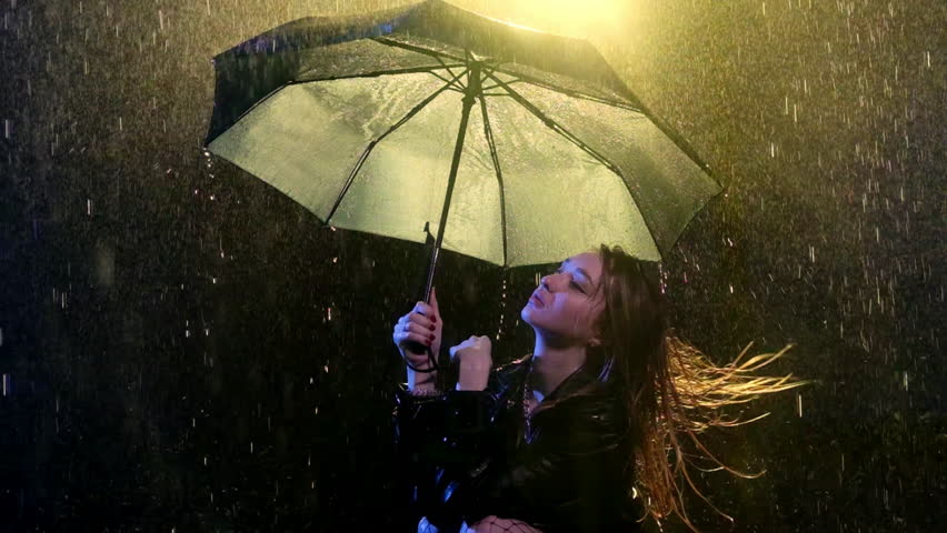 Woman runs through wet hair with hand standing under umbrella. Light of spotlight romanticizes scenery with lady in wet clothes in night rain