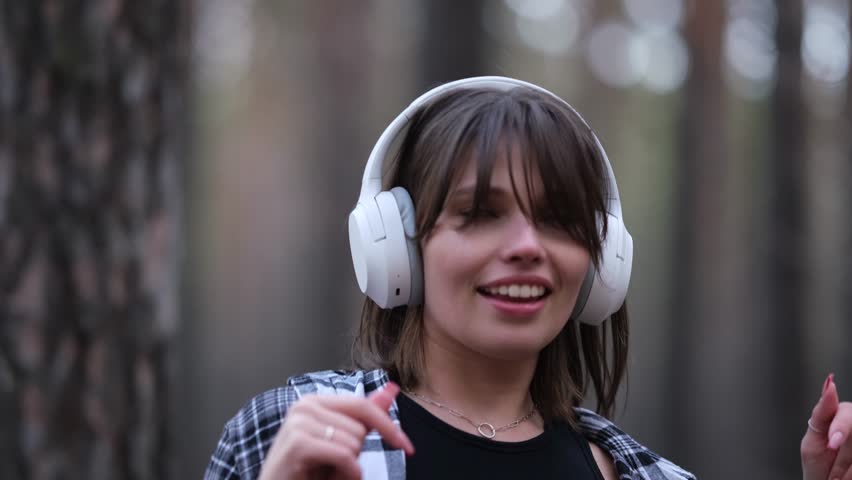 Young beautiful brunette listens to music on headphones outdoors. A woman dances joyfully to music among the maidens in the forest.