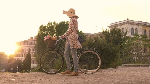 Young woman with bycicle takes pictures of the colosseum in rome at sunset with smartphone. Stylish dress with large hat, flowers and bread in basket.