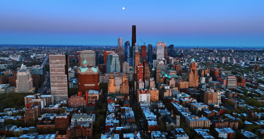 The tiny circle of Moon in the blue sky over the vast scenery of New York, the USA. Drone footage above the metropolis at dusk.