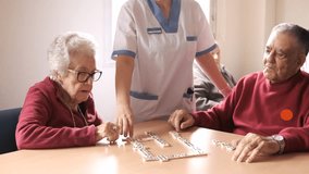 Nurse playing dominoes with senior man and woman in canteen of nursing home