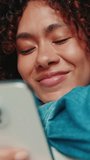 Vertical video, Close-up of smiling young woman with curly hair wearing denim shirt using mobile phone while lying on sofa in cozy living room